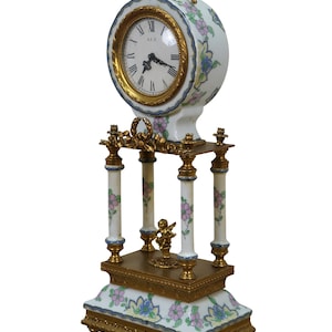 May include: A decorative antique clock with a white floral design and gold accents. The clock face has Roman numerals and gold trim. The clock sits on a gold-colored base with a small cherub statue.
