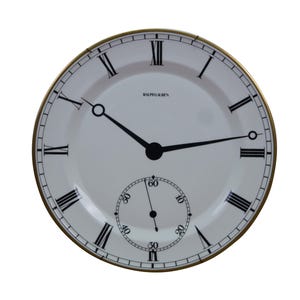 May include: A round wall clock with a white face, black Roman numerals, and a thin gold-toned metal rim. The center features black hands and the text Ralph Lauren. A smaller circular sub-dial at the bottom displays seconds with black numerals.