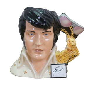 May include: A ceramic mug shaped like the head and shoulders of Elvis Presley, featuring black hair, a high-collared white shirt with gold star accents, and a handle designed as a hand holding sunglasses and a gold lion pendant. Includes an Elvis tag.