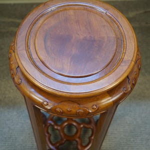 May include: A round, wooden plant stand with a dark brown finish. The top is circular with a raised edge. The stand has a decorative, openwork design near the base and is supported by three legs. The stand is approximately 18 inches tall.