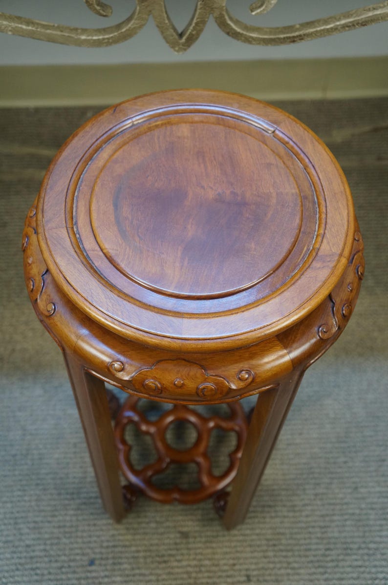 May include: A round, wooden plant stand with a dark brown finish. The top is circular with a raised edge. The stand has a decorative, openwork design near the base and is supported by three legs. The stand is approximately 18 inches tall.