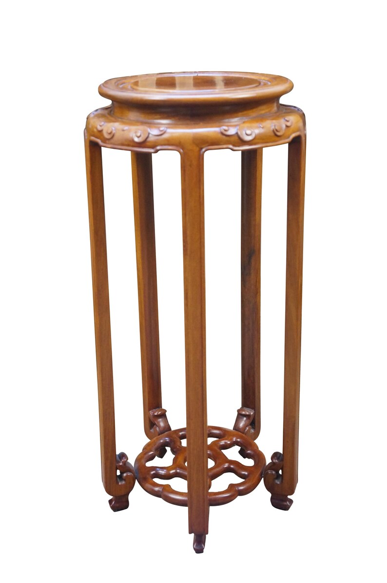 May include: A tall, wooden plant stand with a round top and a decorative, openwork base. The stand is made of a warm-toned wood and features a series of vertical supports. The top has a slightly raised edge. The stand is approximately 30 inches tall.