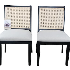 May include: Two black dining chairs with woven cane backs and light gray upholstered seats. The chairs have a classic design, suitable for a dining room or kitchen. One chair has a tag attached.