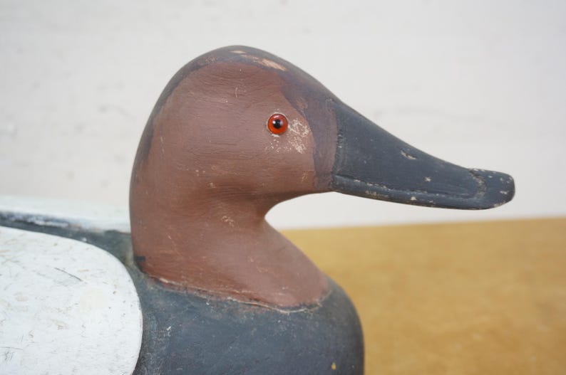 May include: A close-up of a wooden duck decoy. The duck has a brown head and neck, a black beak, and a red eye. The body is painted white and black. The decoy is likely used for hunting or decoration.