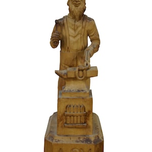 May include: A wooden sculpture of a blacksmith at work. The figure, with a long beard and hat, stands behind an anvil, holding a hammer and a metal piece. The base is inscribed with "ASM".