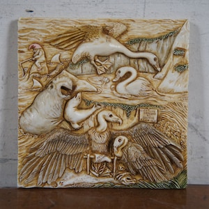 Op de afbeelding: Een witte keramische wandplaat met een reli&euml;fsculptuur van verschillende dieren, waaronder een zwaan, een gans, een zeehond en een gier. De plaat heeft een bruine en groene achtergrond en bevat de tekst "Little Boy Blue, Come Blow Your Horn".