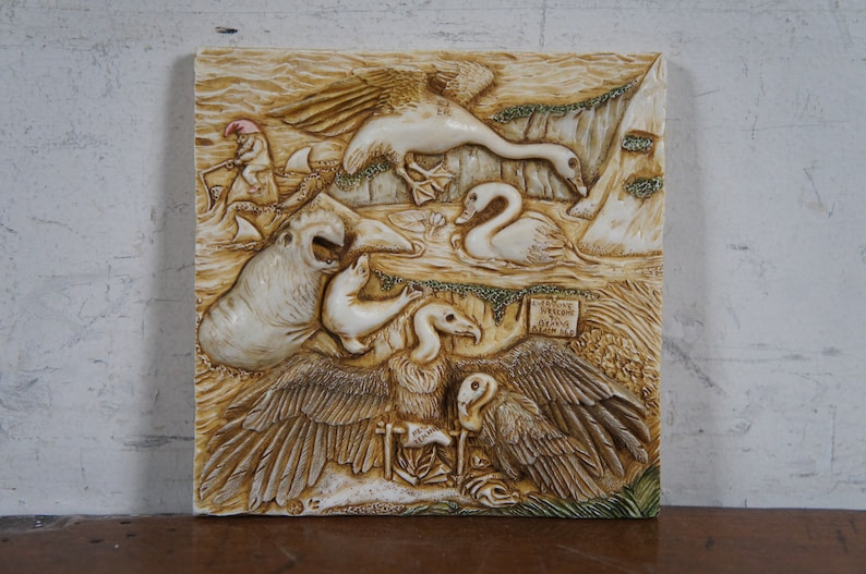 Op de afbeelding: Een witte keramische wandplaat met een reli&euml;fsculptuur van verschillende dieren, waaronder een zwaan, een gans, een zeehond en een gier. De plaat heeft een bruine en groene achtergrond en bevat de tekst "Little Boy Blue, Come Blow Your Horn".