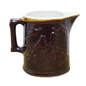 May include: A dark brown ceramic creamer pitcher featuring a glossy finish and a white interior. The exterior surface displays a raised relief design of two figures standing amidst foliage, with a textured border along the rim and base.