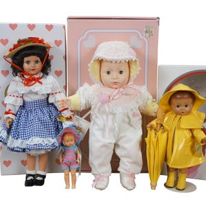 May include: Four vintage dolls are displayed in front of their boxes. One doll wears a blue gingham dress and a straw hat. Another doll is dressed in a white outfit and bonnet. A third doll wears a yellow raincoat and holds an umbrella. A small doll is also present.