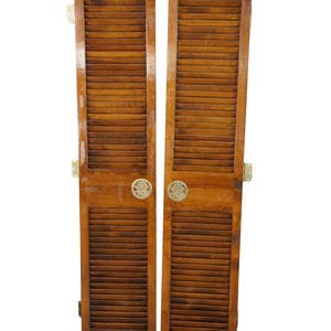 May include: A pair of tall, rectangular wooden shutters with a medium brown finish. Each shutter features horizontal slats in the upper and lower sections, a solid middle rail, and a circular brass pull with an engraved character design.