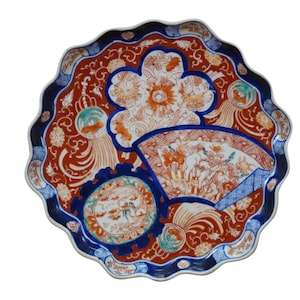 May include: A decorative ceramic plate with a scalloped edge. The plate features a vibrant design with a red background, blue accents, and orange and white floral and figurative motifs. The plate's design includes a fan and circular elements.
