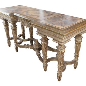 May include: A long, ornately carved wooden console table with a gold finish. The table has a rectangular top with a decorative inlay and four legs with intricate details.