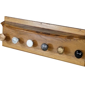 May include: A rectangular wooden wall rack featuring a top shelf and six distinct knobs. The knobs vary in appearance, including a metallic gold, a clear glass, a solid white, a dark textured metal, a gold textured metal, and a solid black knob.