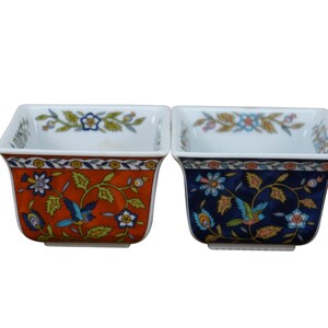 May include: Two square ceramic bowls with floral and bird designs. One bowl is orange with blue birds and green leaves, the other is navy blue with similar imagery. Both have white interiors and floral patterns on the rims.