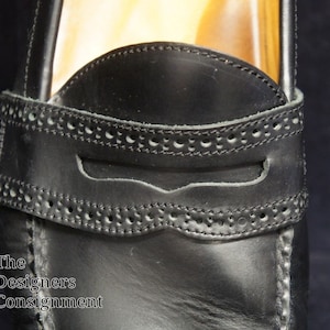 May include: Close-up of a black leather loafer with a decorative stitch pattern around the opening. The loafer has a leather strap with a small opening.