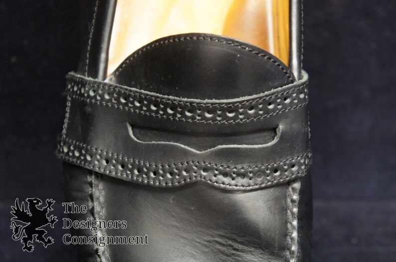 May include: Close-up of a black leather loafer with a decorative stitch pattern around the opening. The loafer has a leather strap with a small opening.