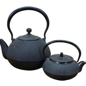 May include: Two black cast iron teapots. The larger teapot has a curved handle and a lid with a small knob. The smaller teapot also has a curved handle and a lid with a knob. Both teapots have a textured surface.