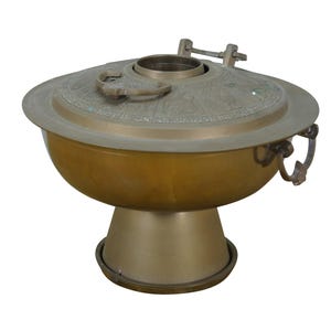 May include: A brass-colored, antique-style container with a lid. The container has a rounded bowl shape, a pedestal base, and a lid with a handle. The lid has a decorative pattern. The container has two handles on the sides.