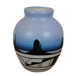 May include: A small, light blue ceramic vase with a white and black geometric design. The vase is painted with a scene of a desert landscape with a blue sky and black mountains.