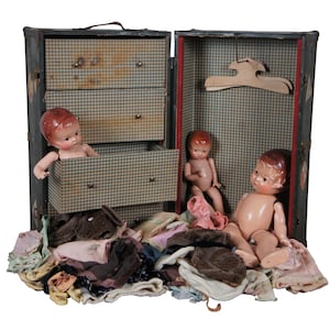 May include: A vintage wooden doll trunk with three drawers and a hinged lid. The trunk is open and contains three dolls, a hanger, and a pile of doll clothes.