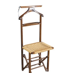 May include: A vintage wooden valet chair with a woven seat. The chair features a built-in clothes hanger and a small shelf. The wood has a warm, brown tone, and the seat is made of a light-colored woven material.