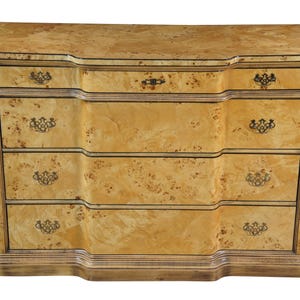 May include: A light-colored wooden dresser with three drawers and a curved front. The dresser has a speckled pattern and ornate drawer pulls. The top of the dresser has a curved edge.