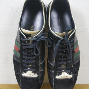 May include: Pair of black sneakers with green and red stripes. The sneakers have black laces and the word "GUCCI" in gold lettering on the tongue. The shoes have a suede toe and a gold trim.