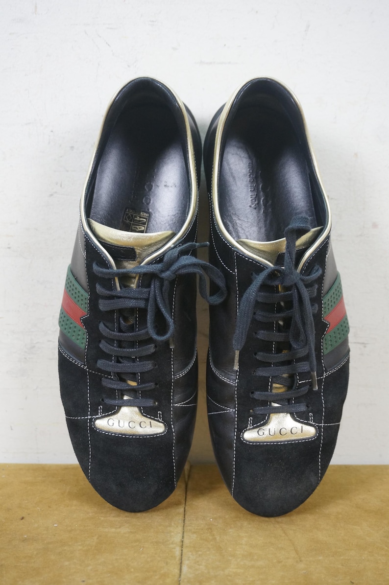 May include: Pair of black sneakers with green and red stripes. The sneakers have black laces and the word "GUCCI" in gold lettering on the tongue. The shoes have a suede toe and a gold trim.