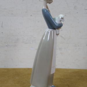 May include: A porcelain figurine of a woman in a long dress, holding a lamb. The woman's dress is gray and blue, with a white apron. The lamb is white. The figurine has a light green base.