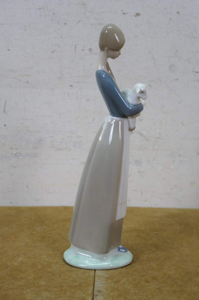 May include: A porcelain figurine of a woman in a long dress, holding a lamb. The woman's dress is gray and blue, with a white apron. The lamb is white. The figurine has a light green base.