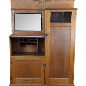 May include: A tall oak wood hall tree featuring a rectangular mirror, a small open shelf with internal dividers, a lower cabinet door, and a tall side cabinet with a glass panel at the top. The piece is topped with a carved wooden shell motif.