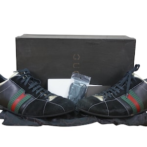 May include: Black and gold sneakers with green and red stripes. The shoes are displayed with a black box and accessories. The sneakers have black laces and a suede and leather design. The brand name is visible on the box.