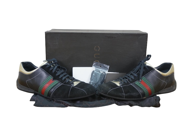 May include: Black and gold sneakers with green and red stripes. The shoes are displayed with a black box and accessories. The sneakers have black laces and a suede and leather design. The brand name is visible on the box.