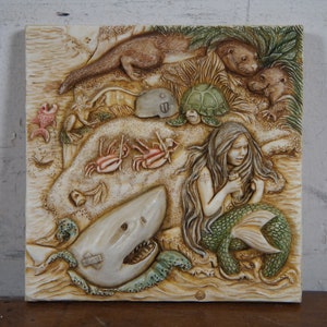 Op de afbeelding: Een witte keramische wandplaat met een reli&euml;fsculptuur van een zeemeermin, een haai, een schildpad, otters en krabben. De zeemeermin houdt een fles in haar hand. De plaat heeft een beige achtergrond.