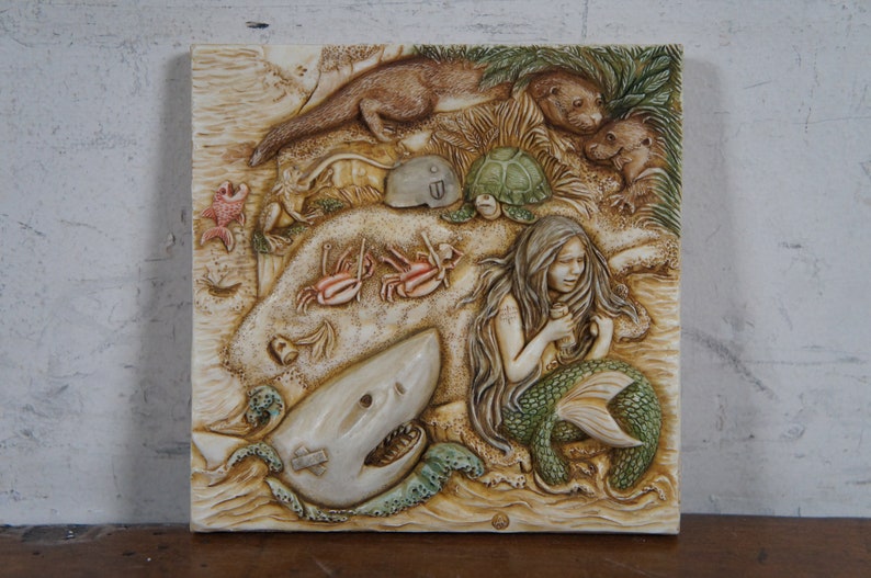Op de afbeelding: Een witte keramische wandplaat met een reli&euml;fsculptuur van een zeemeermin, een haai, een schildpad, otters en krabben. De zeemeermin houdt een fles in haar hand. De plaat heeft een beige achtergrond.