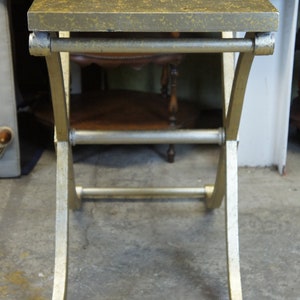 K&ouml;nnte beinhalten: Ein goldfarbener Beistelltisch aus Metall mit einer patinierten goldenen Tischplatte. Der Tisch hat ein schlichtes, modernes Design mit einem geschwungenen, X-f&ouml;rmigen Gestell.