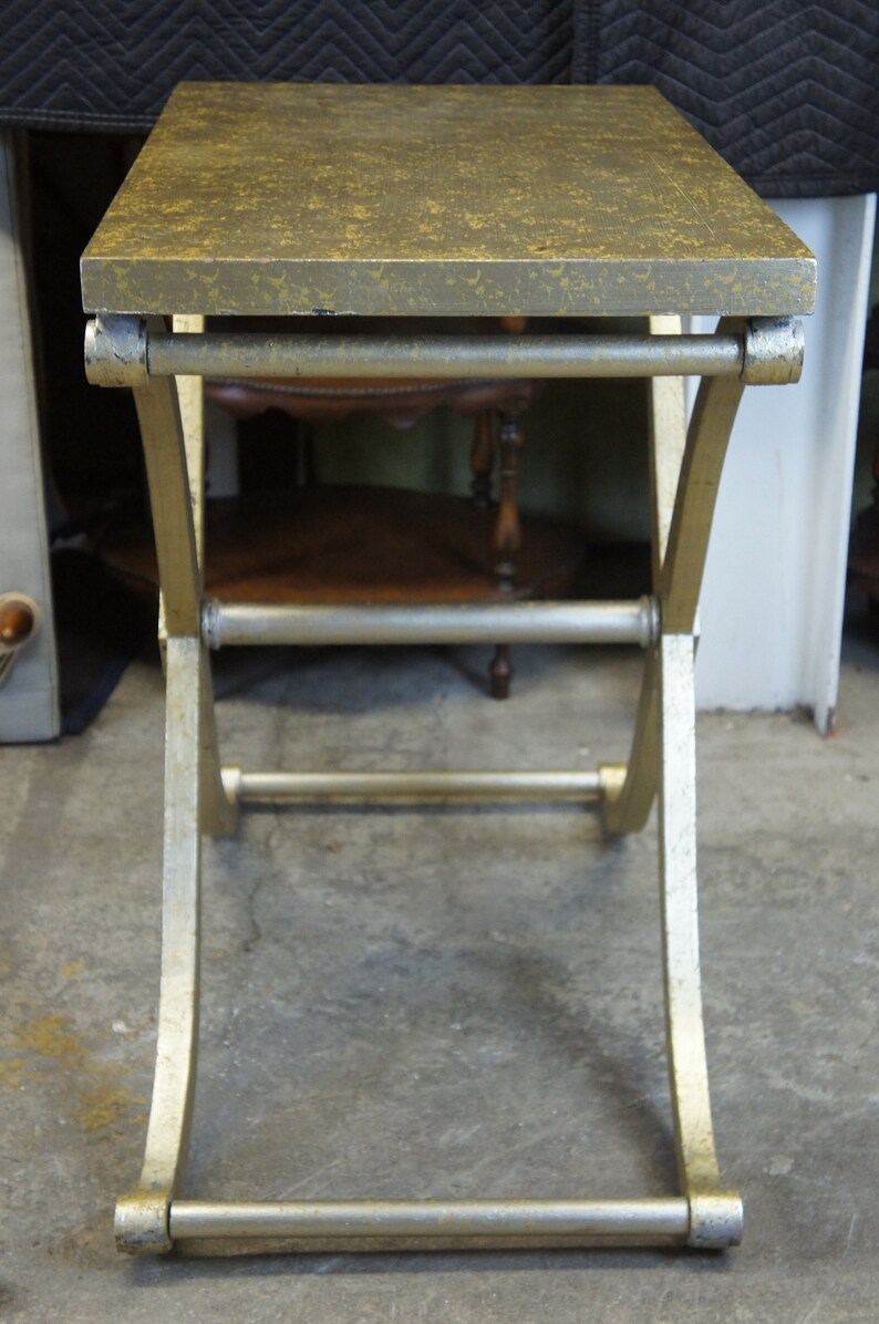 K&ouml;nnte beinhalten: Ein goldfarbener Beistelltisch aus Metall mit einer patinierten goldenen Tischplatte. Der Tisch hat ein schlichtes, modernes Design mit einem geschwungenen, X-f&ouml;rmigen Gestell.