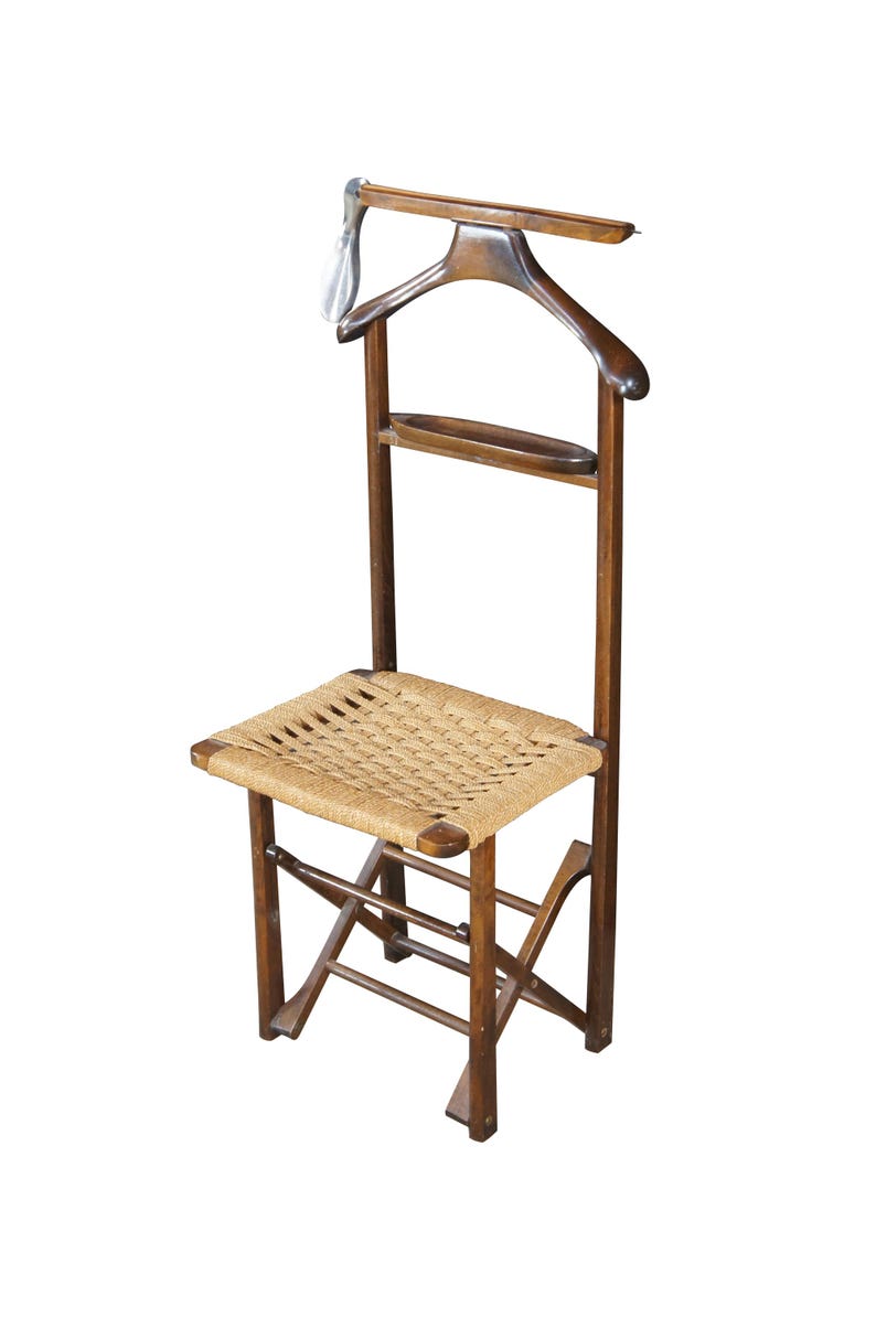 May include: A vintage wooden valet chair with a woven seat. The chair features a built-in hanger and a small shelf. The wood has a warm, brown tone, and the seat is made of a light-colored woven material.