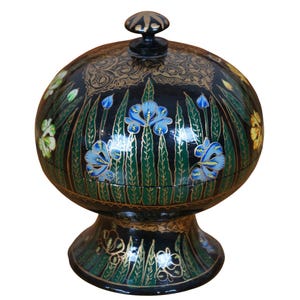 May include: A black and gold painted wooden box with a round lid and a base. The box is decorated with green leaves and blue flowers.