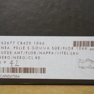 May include: A close-up of a label on a black box. The label includes text such as "SNEA. PELLE S.GOMMA SUE/FLOR" and "NERO/NERO/CL.RE". The size is indicated as 42. A barcode and a shoe illustration are also present.