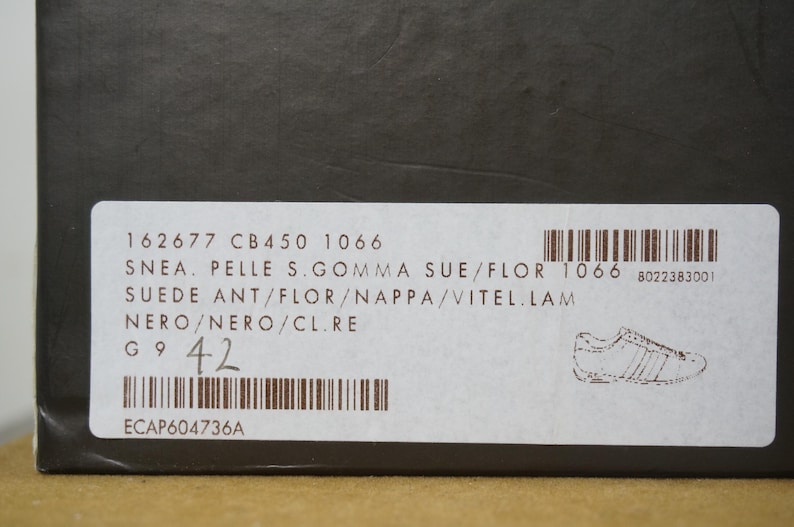 May include: A close-up of a label on a black box. The label includes text such as "SNEA. PELLE S.GOMMA SUE/FLOR" and "NERO/NERO/CL.RE". The size is indicated as 42. A barcode and a shoe illustration are also present.