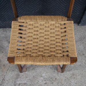 May include: A woven rope seat chair with a dark wooden frame. The seat is made of tightly woven, light brown rope, creating a textured surface. The chair's legs are angled, providing stability. The chair is a vintage piece.