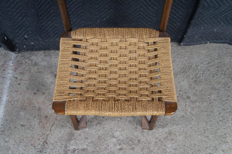 May include: A woven rope seat chair with a dark wooden frame. The seat is made of tightly woven, light brown rope, creating a textured surface. The chair's legs are angled, providing stability. The chair is a vintage piece.