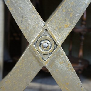 K&ouml;nnte beinhalten: Nahaufnahme eines goldfarbenen, X-f&ouml;rmigen Metallrahmens mit einem rautenf&ouml;rmigen Design in der Mitte. Die Raute hat ein kreisf&ouml;rmiges Design mit einem wei&szlig;en Knopf in der Mitte.