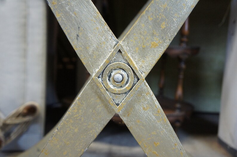 K&ouml;nnte beinhalten: Nahaufnahme eines goldfarbenen, X-f&ouml;rmigen Metallrahmens mit einem rautenf&ouml;rmigen Design in der Mitte. Die Raute hat ein kreisf&ouml;rmiges Design mit einem wei&szlig;en Knopf in der Mitte.