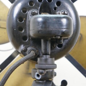 Puede incluir: Primer plano de un ventilador el&eacute;ctrico antiguo. La carcasa del motor del ventilador es negra con varios orificios de ventilaci&oacute;n. Las aspas son de color dorado. Un cable de alimentaci&oacute;n negro sale del motor.