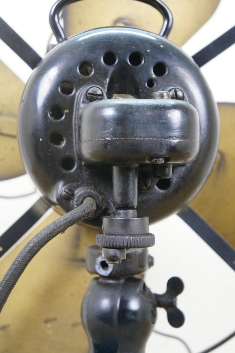 Puede incluir: Primer plano de un ventilador el&eacute;ctrico antiguo. La carcasa del motor del ventilador es negra con varios orificios de ventilaci&oacute;n. Las aspas son de color dorado. Un cable de alimentaci&oacute;n negro sale del motor.
