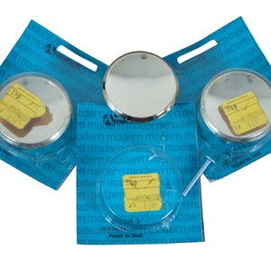 May include: Three round, silver-colored, decorative mirror tiles packaged in blue plastic packaging with the text "Amerock Modern Modern" printed on the front. Each tile has a yellow price tag with the text "300" printed on it.