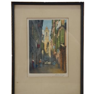 May include: Framed print of a European street scene. The artwork depicts a narrow street with buildings on either side, featuring green shutters and laundry hanging from windows. A church tower is visible in the distance. The print is in a dark wooden frame.