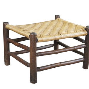 May include: A small, square footstool with a woven, light-colored seat. The frame is made of dark brown wood, with four sturdy legs and horizontal supports. The footstool has a rustic, handcrafted appearance.